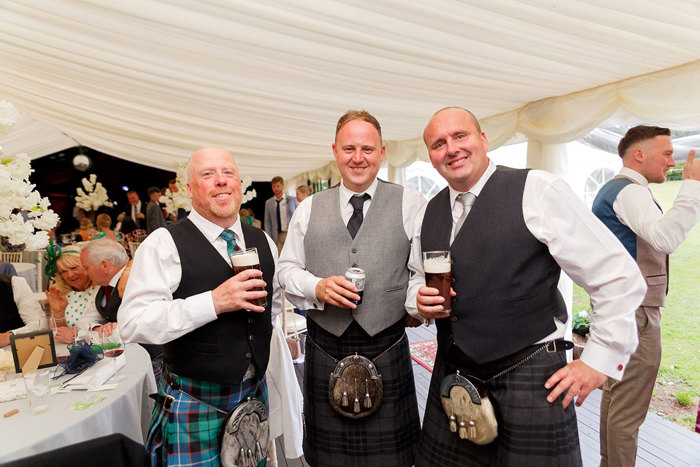 Three men in kilts each holding a glass of beer look at the camera