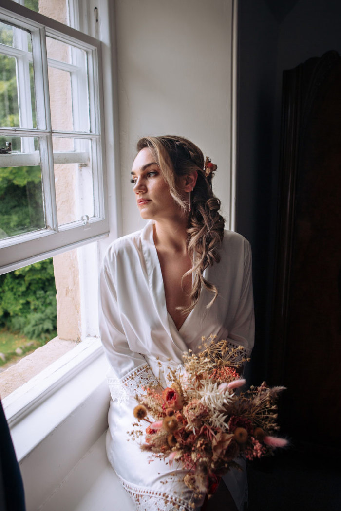 woman sits on a windowsill in a silky white robe and her hair styled in loose curls while holding a dried flower arrangement