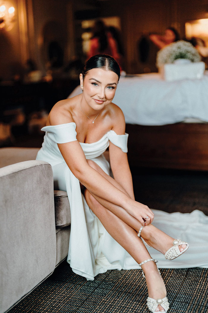 bride in strapless wedding dress with off the shoulder straps bends down to fasten strap on her pearl heels