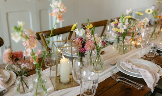 bud vases and candles line the middle of white table runner on a wooden dining table