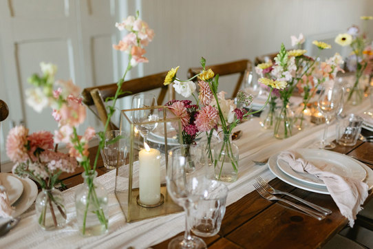 bud vases and candles line the middle of white table runner on a wooden dining table