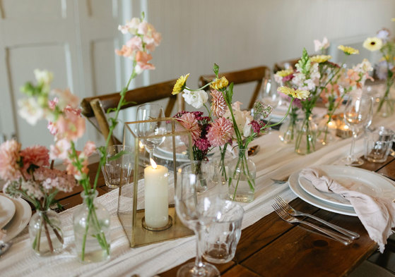 bud vases and candles line the middle of white table runner on a wooden dining table