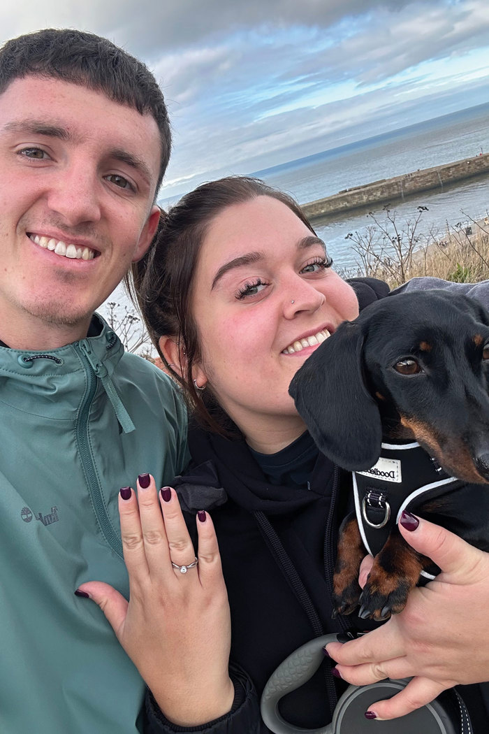 a man and a woman posing for a selfie holding a dachshund dog in front of a body of water