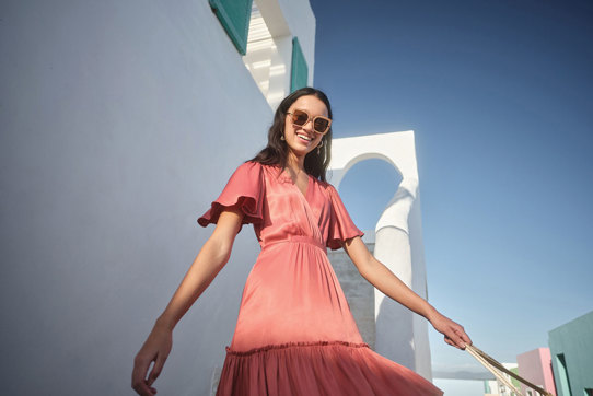 girl smiling wearing a coral dress with floaty sleeves in front of a white wall in the sunshine