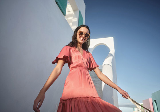 girl smiling wearing a coral dress with floaty sleeves in front of a white wall in the sunshine