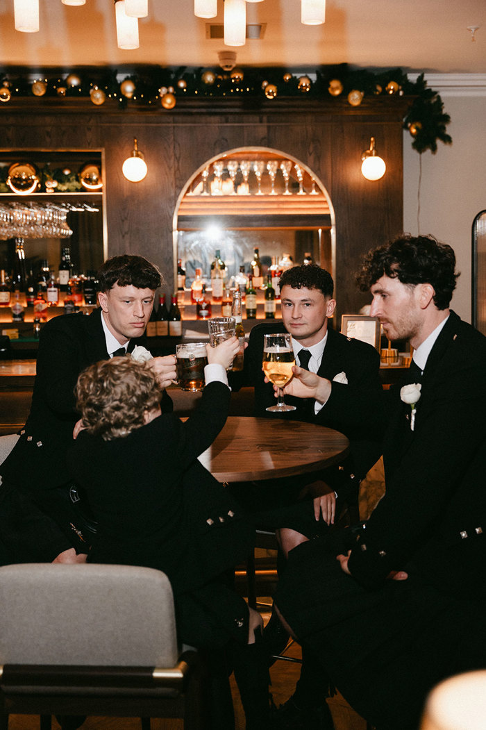 three men and one young boy sit around a round table in a bar setting, each holding their own drink up and out to cheers with the rest