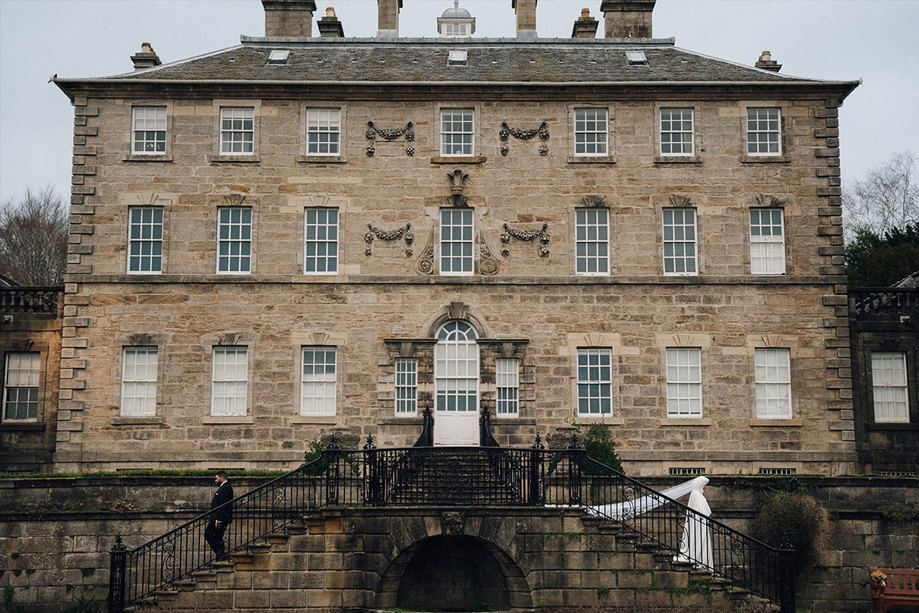 A large country house with two sets of stairs leading away from the main door, a bride walks down one set of stairs and a groom walks down the other