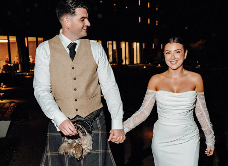 bride in strapless dress with detached tulle sleeves holds hand of tall groom in beige and dark green kilt outfit