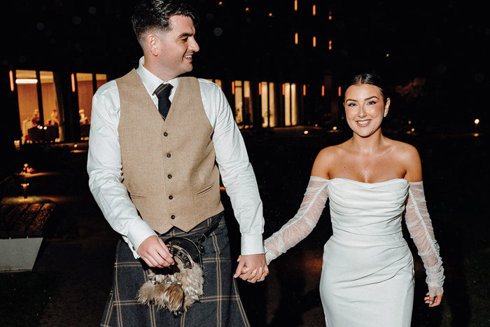 bride in strapless dress with detached tulle sleeves holds hand of tall groom in beige and dark green kilt outfit