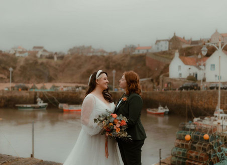 two brides, one wearing a wedding dress, one wearing a suit, look into each others' eyes with Crail and harbour in the background