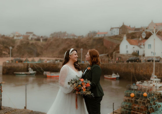 two brides, one wearing a wedding dress, one wearing a suit, look into each others' eyes with Crail and harbour in the background