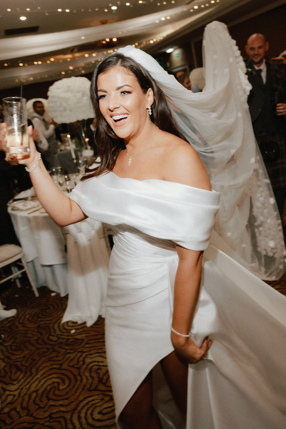 bride with dark hair in off the shoulder wedding dress with a leg slit smiles and holds out a drink while a man in the background holds her long veil out behind her