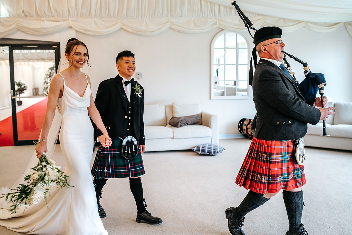 bride and groom hold hands walking into marquee at mar hall as piper wearing red tartan kilt leads them in