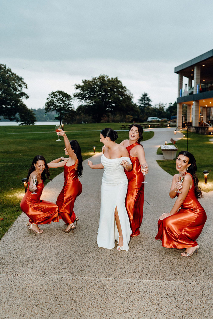 bride in strapless wedding dress with off the shoulder straps stands outside on a paved path with four bridesmaids in matching burnt orange dresses holding glasses of champagne