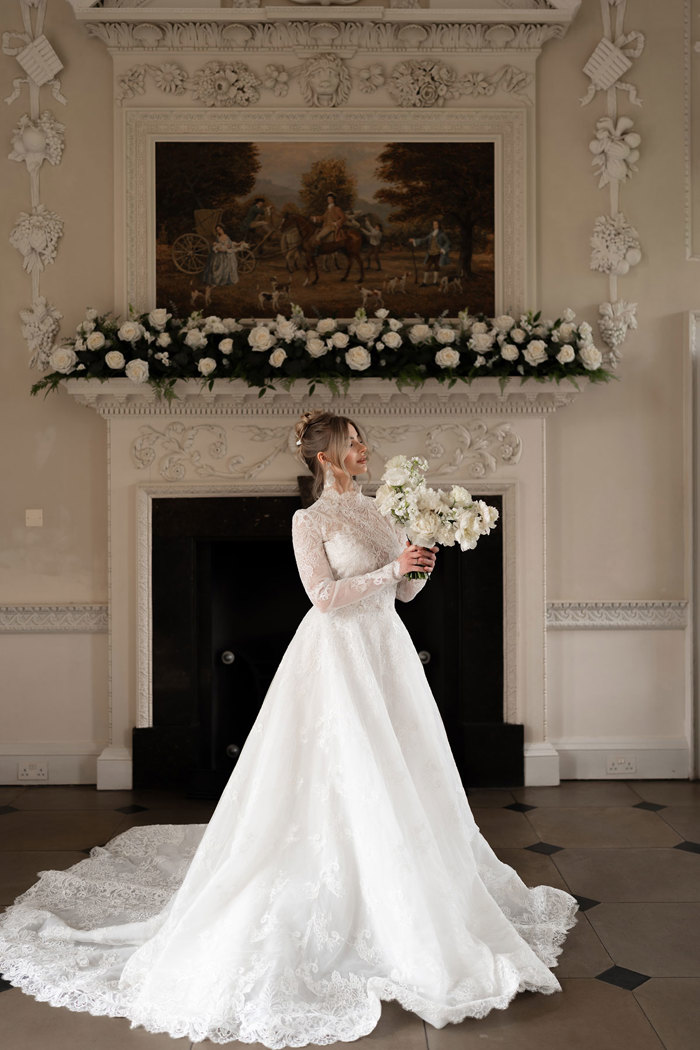 woman in high neck long lace sleeve wedding dress holds a bouquet of flowers as she stands in front of unused fireplace with garland display of white roses