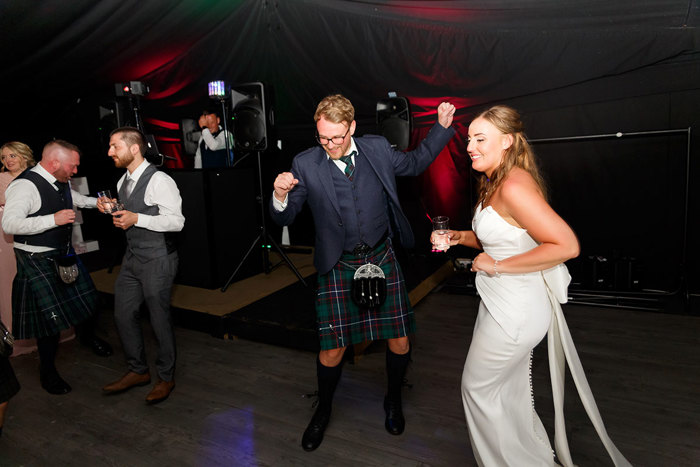 Men in suits and kilts and a bride in a white dress dancing 