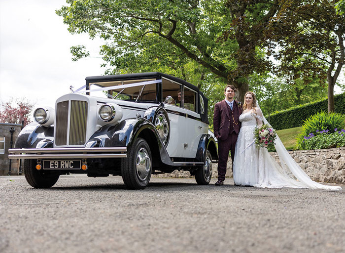 A bride in a white dress and a groom in a brown suit standing next to a vintage white wedding car