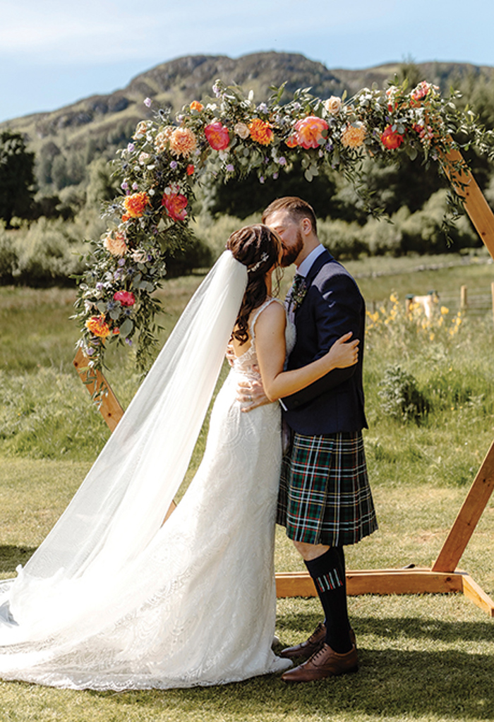 bride and groom kissing in front of floral arch with green hills and blue skies in the background