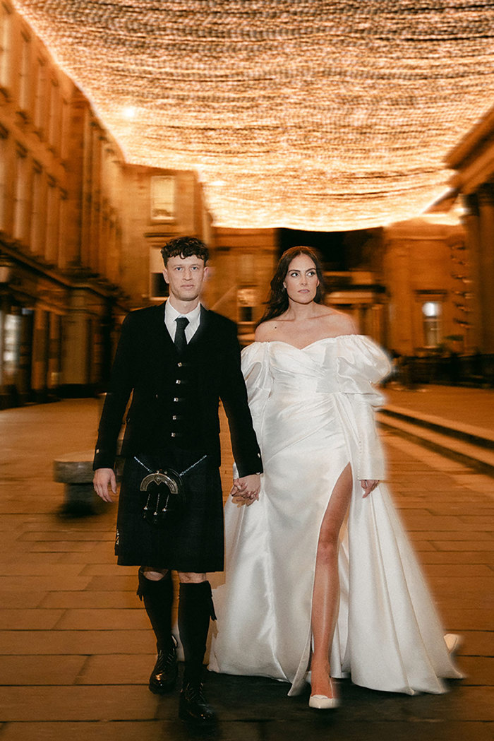 brunette bride in high leg slit wedding dress with long puff sleeves and groom in full dark tartan kilt outfit walk hand in hand outdoors with a canopy of twinkling lights in the night sky above them