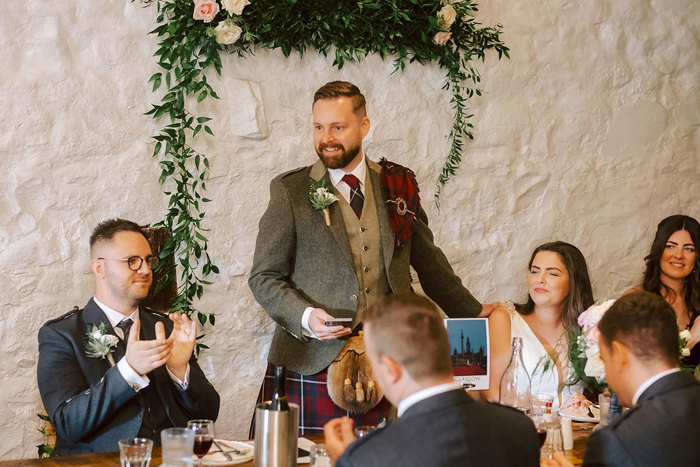 A groom standing to give a speech at a wedding as the guests looking on clap 