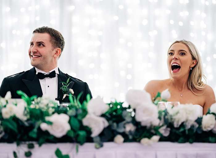 a surprised looking bride and groom pulling a funny expression sitting at a white table decorated with white roses and greenery