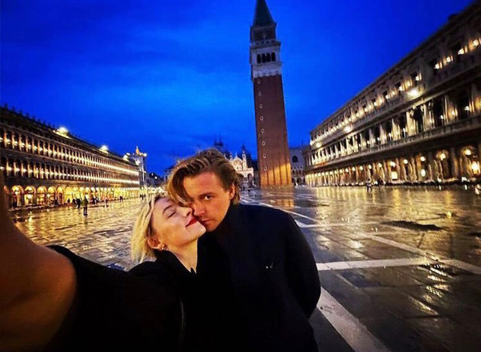 A woman takes a selfie with her arm stretched out next to the camera as she cuddles up to a man in a town square