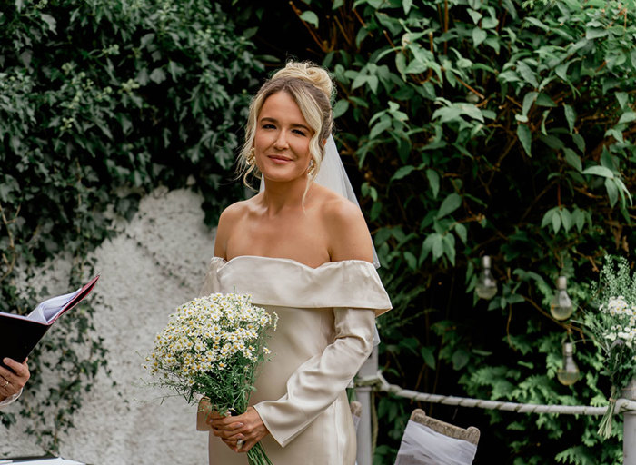 a bride wearing an off-shoulder long-sleeve ivory wedding dress holding a large bunch of daisies with greenery and white pebble-dash wall in background