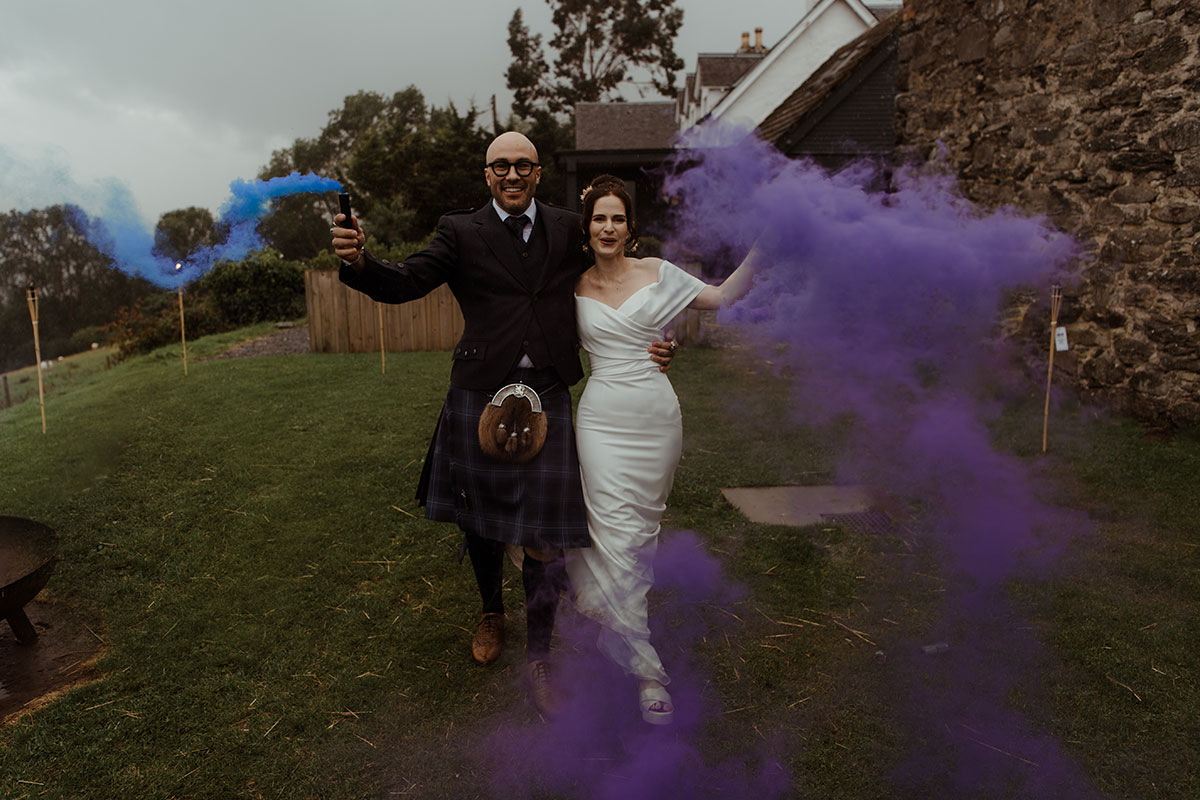 groom in blue, grey and white tartan kilt outfit holds out a blue smoke stick to one side while bride to his other side, whom he has an arm around, holds out a purple smoke stick to her other side