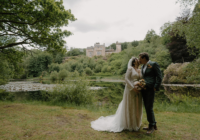 bride and groom kissing while stood outdoors in grassy area with large brown building in background