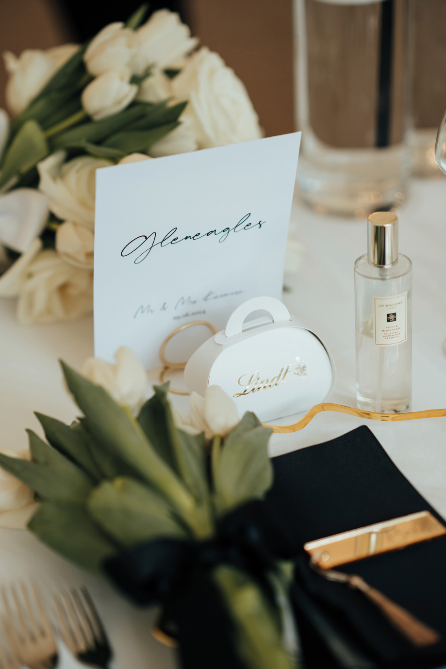 A bouquet of white roses sits next to a black napkin, cutlery, a bottle of Jo Malone perfume and a white Lindt chocolate box