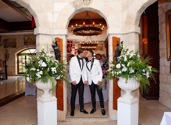 two men kissing below a narrow stone arch in an old stone building. There are large white flower arrangements in urns on either side of them in the foreground