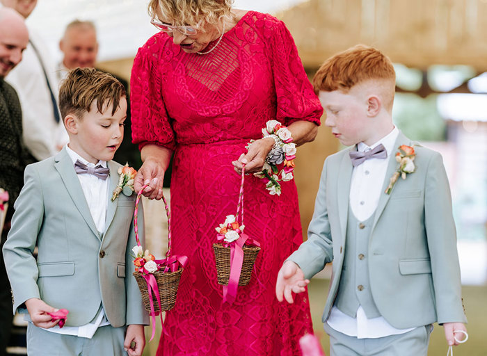 an older lady wearing a hot pink dress holding two small wicker baskets decorated with ribbon. Two young boys wearing powder blue suits stand either side of her