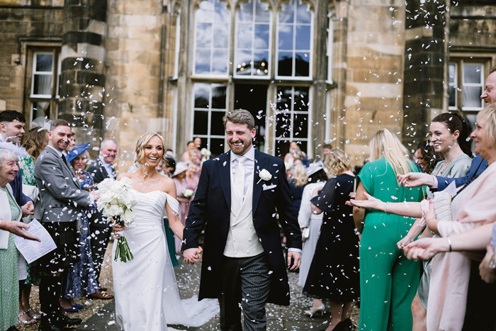 A bride and groom holding hands and walking outside Mar Hall with confetti falling.