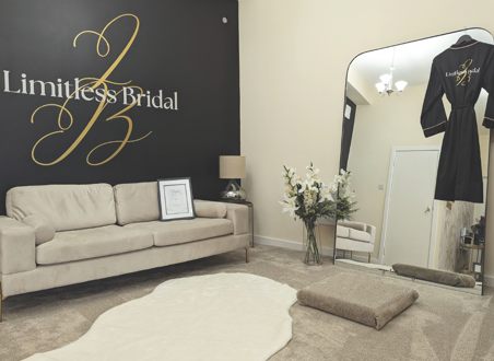 dressing room with beige carpet and couch, with flowers reflected in mirror that has black robe hanging on it. 'Limitless Bridal' is on the wall in gold and white writing. 