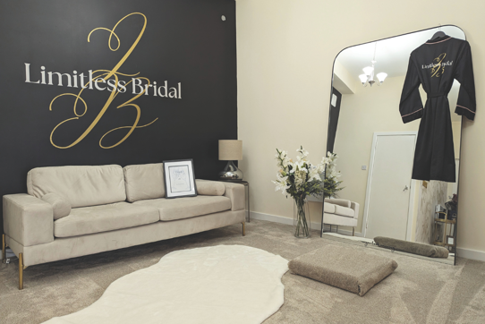 dressing room with beige carpet and couch, with flowers reflected in mirror that has black robe hanging on it. 'Limitless Bridal' is on the wall in gold and white writing. 