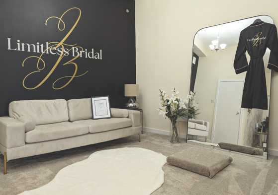 dressing room with beige carpet and couch, with flowers reflected in mirror that has black robe hanging on it. 'Limitless Bridal' is on the wall in gold and white writing. 