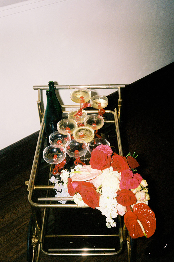 a gold cocktail cart and an assortment of empty and filled champagne coupe glasses beside two popped champagne bottles and a a pink and red flower bouquet