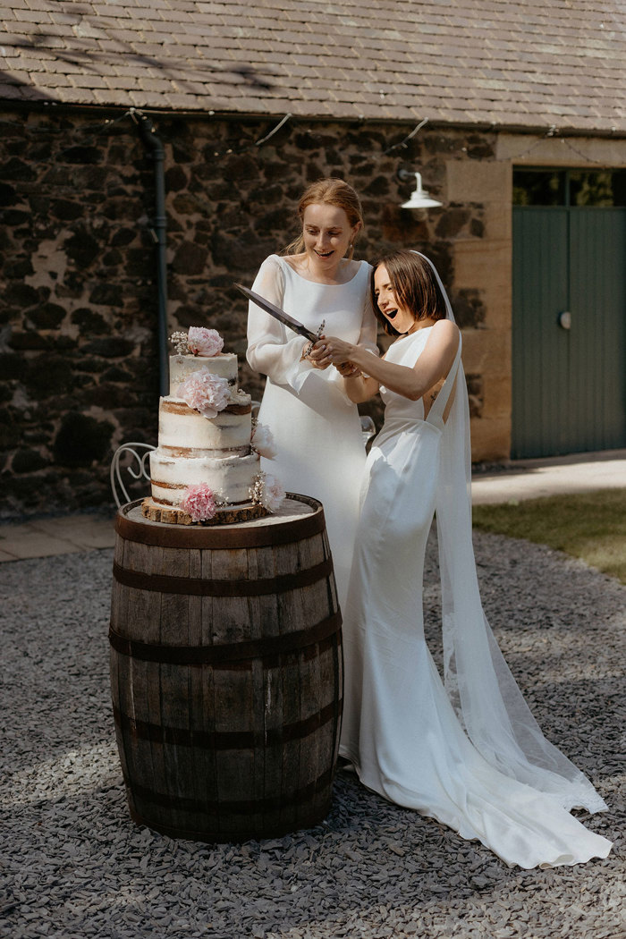 two brides cutting a semi naked buttercream wedding cake sitting on a wooden barrell with a sword in a stone courtyard setting