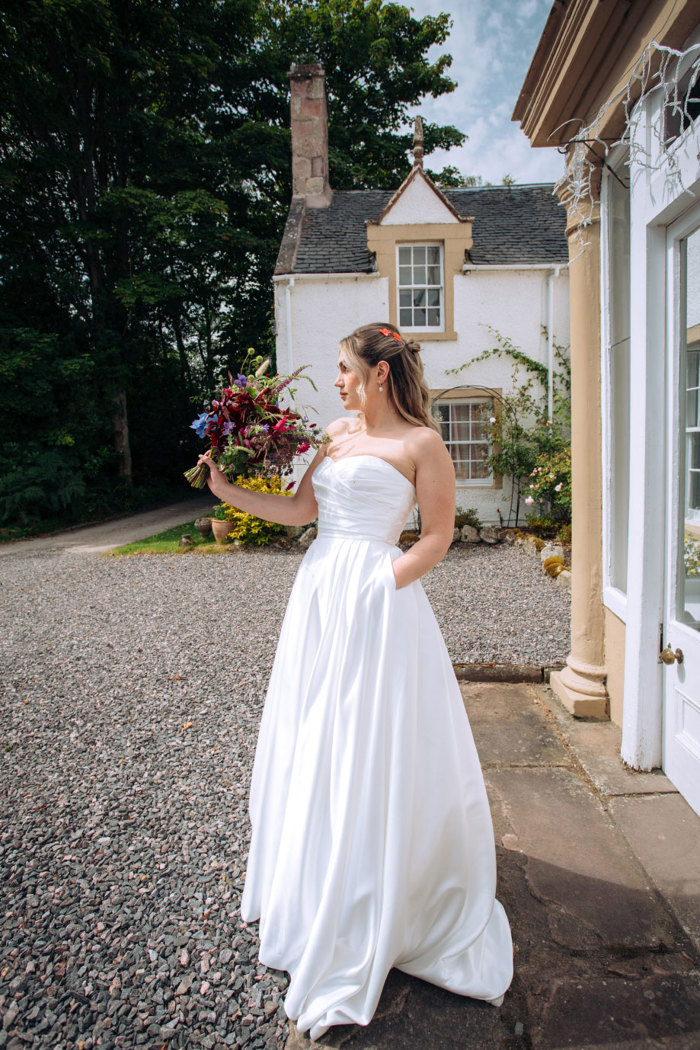 bride in strapless wedding dress stands outdoors with a bouquet held up in one hand and the other hand in her pocket