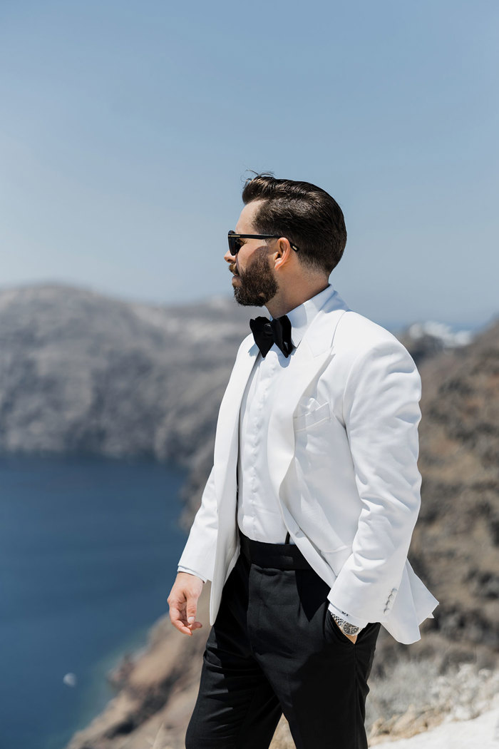 dark haired man wears sunglasses and a white jacket tuxedo as he looks out at a bright blue ocean from white clifftop