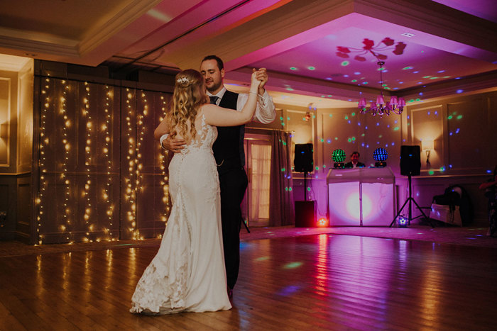 A bride faces away from the camera as she dances with the groom who looks down at her as a colourful DJ booth has lights shining from it behind them