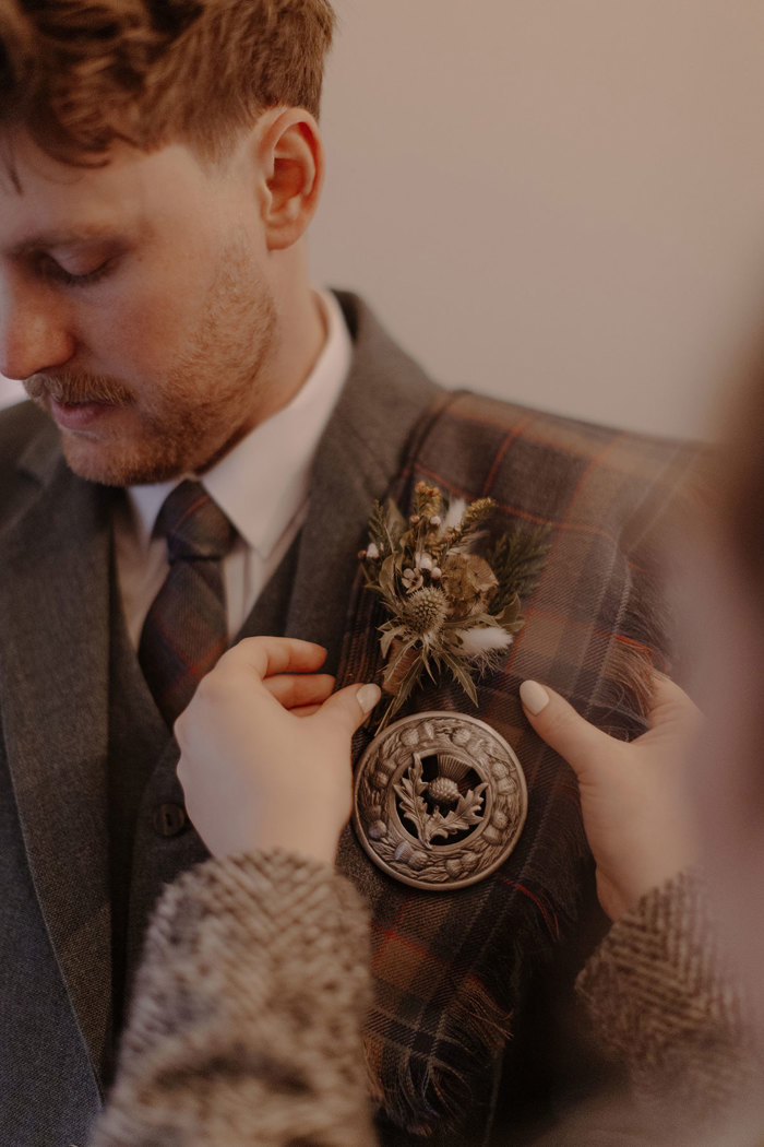 a pair of manicured hands straighten out a man's tartan fly plaid, kilt pin and dried flower boutonnière