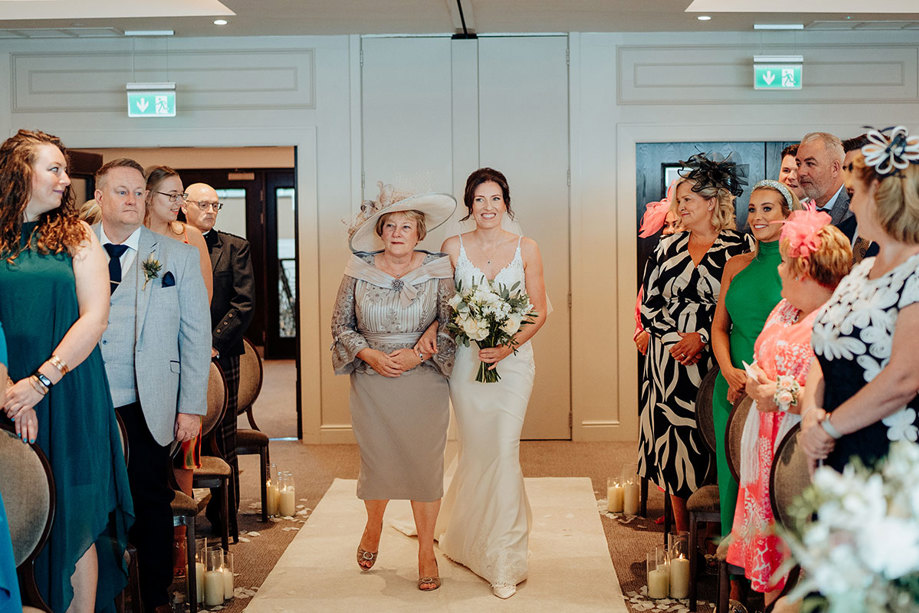 a bride walks down a white carpet on her wedding day at Seamill Hydro on the arm of her mum as guests smile and look on