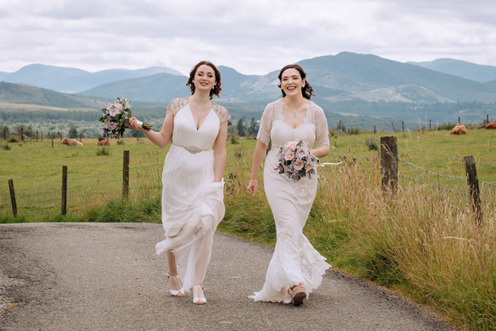 Two Brides Carrying Bouquets Walking Beside A Field