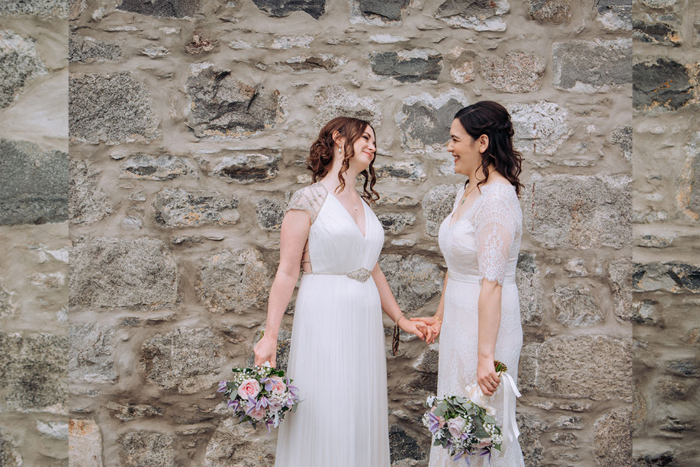 Two Smiling Brides Standing And Holding Hands Against A Stone Wall