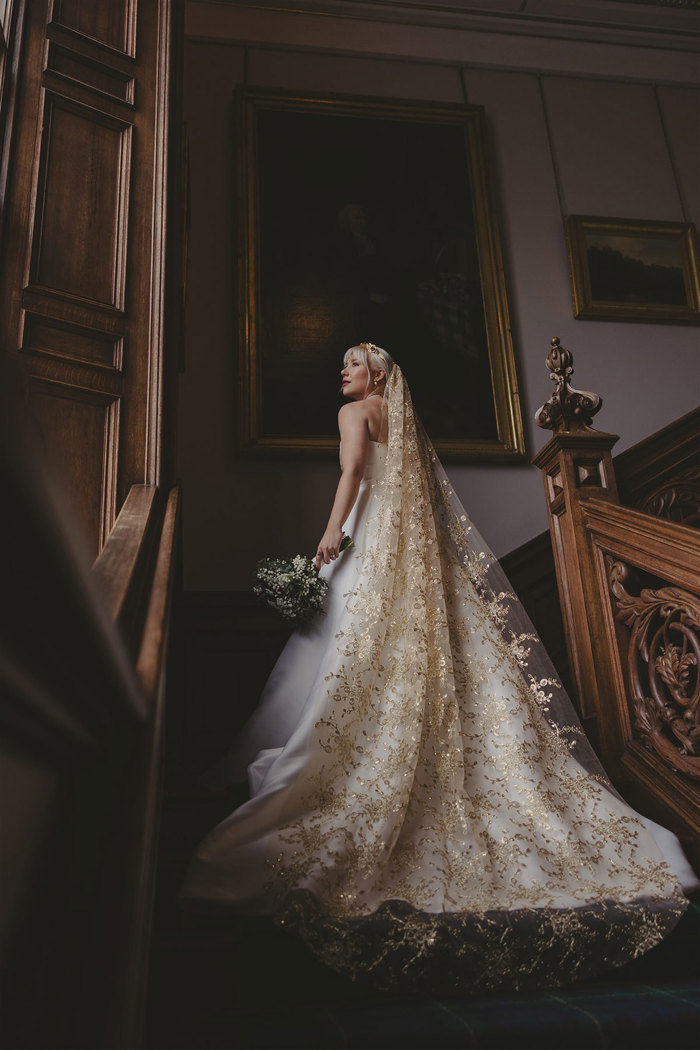 A bride standing on a wooden staircase. She is wearing a gold embroidered veil.