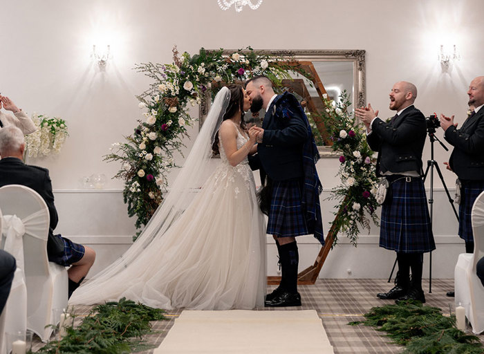 a bride and groom kissing in front of a hexagonal wooden arch decorated with flowers during a wedding ceremony at Nethybridge Hotel. Other guests are clapping and cheering