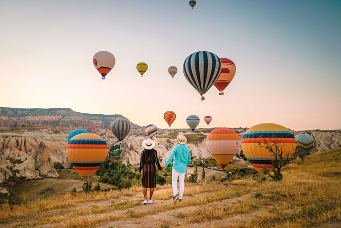 couple holding hands and looking up at a sky filled with hot air balloons