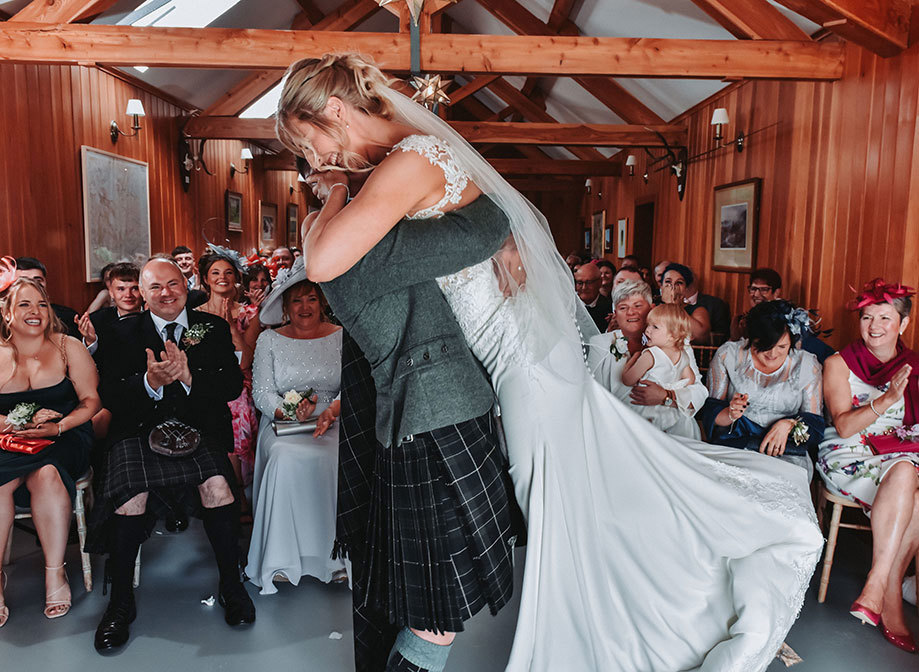 a groom lifting a bride up as rows of seated wedding guests behind them cheer and laugh. They are in a wooden clad building