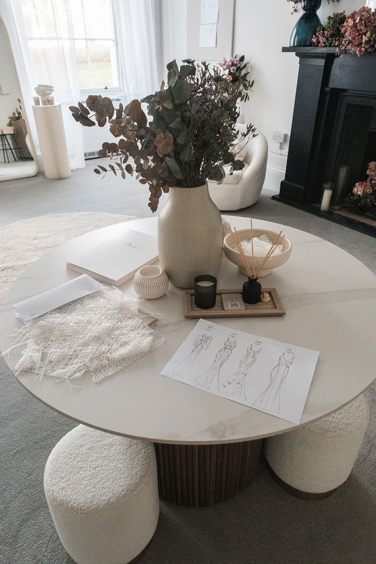 round marble table with soft cream stools gathered around has a vase of flowers in the middle and fabric samples and dress sketches laid out on top